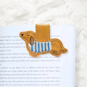 Könnte beinhalten: Ein hellbrauner Leder-Lesezeichen in Form eines Dackelhundes. Der Hund hat braune Stickdetails und einen blau-weiß gestreiften Pullover. Das Lesezeichen befindet sich auf den Seiten eines Buches.