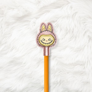 May include: A pink embroidered pencil topper featuring a cartoon character in a bunny costume. The character has a yellow face with a wide grin and pointed ears. The pencil is orange.