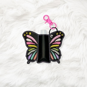 May include: Black butterfly-shaped hand sanitizer holder with colorful embroidered wings. The holder has a pink clip and a black ring for attachment. The wings feature pink, yellow, and blue accents. The holder contains a small, clear bottle.