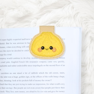 May include: A yellow felt dumpling bookmark with a smiling face and blush accents. The bookmark has a tan leather tab and is placed on a book. The dumpling design is a cute and whimsical accessory for book lovers.