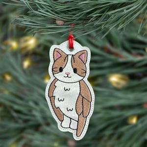 May include: Embroidered cat ornament with a white body, brown and tan markings, and a red hanging loop. The cat is smiling and has a cute design. The ornament is hanging on a Christmas tree.