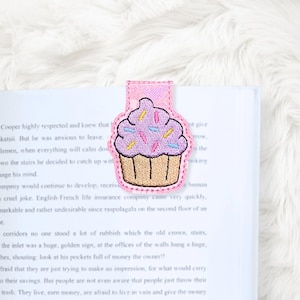 May include: A pink cupcake-shaped bookmark with embroidered details. The bookmark features a light purple frosting with colorful sprinkles and a tan cupcake base. It is attached to the top of a book.