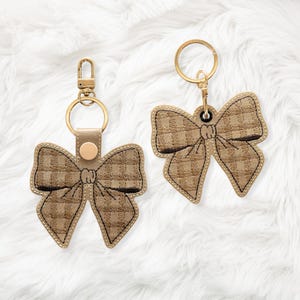 May include: Two bow-shaped keychains. The bows are tan with a plaid pattern and black trim. The keychains have gold-colored hardware, including a clasp and a ring. The background is a soft, white, fluffy material.