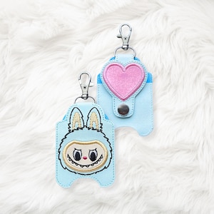 May include: Two light blue keychains with silver-colored clasps. One keychain features a cartoon monster face with a furry blue body and a smiling mouth. The other keychain has a pink heart design.