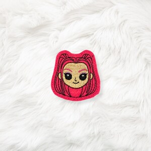 May include: A red embroidered patch featuring a cartoon girl with pink hair and a smiling face. The patch has a gold skin tone and black details. The patch is on a white, fluffy surface.