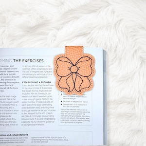 May include: A peach-colored leather bookmark shaped like a bow, with black stitching outlining the design. The bookmark is resting on a book with text about exercises.