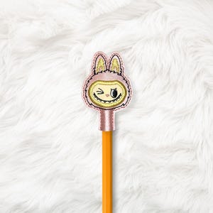 May include: A pink and gold embroidered pencil topper featuring a cartoon character with a winking eye and a mischievous grin. The topper is attached to an orange pencil, set against a fluffy white background.