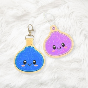 May include: Two embroidered fig-shaped keychains with smiling faces. One is blue with a metal lobster clasp, and the other is purple with a silver ball chain. Both feature light-colored stitched borders and pink blush details on the cheeks.
