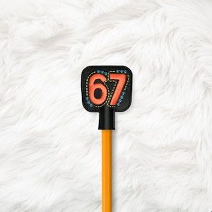 May include: A black leather pencil topper embroidered with the number 67 in orange thread. The number is outlined in yellow stitching and surrounded by blue decorative stitching. The pencil is orange.
