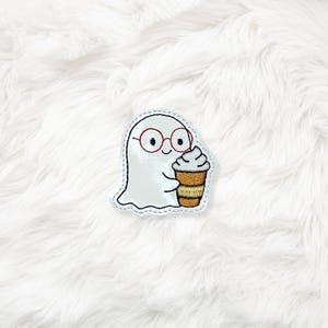 以下が含まれることがあります： 赤いメガネをかけた、ホイップクリーム付きのコーヒーを持った、フレンドリーなゴーストの刺繍パッチ。ゴーストは白で、水色の輪郭があります。コーヒーカップは茶色とベージュです。