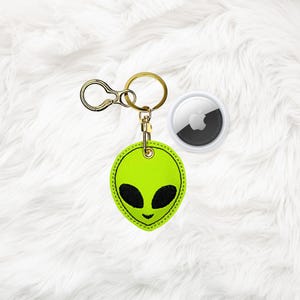 May include: A neon green alien face keychain with black eyes and a smiling mouth. The keychain has a gold-colored clasp and ring. An Apple AirTag is next to the keychain.