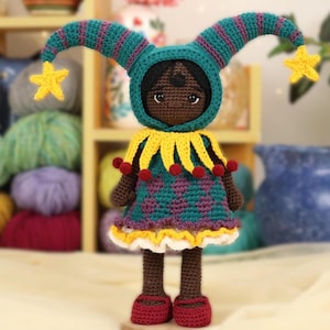 Puede incluir: Una muñeca de crochet que lleva un traje de bufón verde y morado con detalles amarillos y rojos. La muñeca tiene la piel morena y los ojos negros.