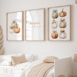 May include: Three framed art prints with a fall theme. The left print features watercolor pumpkins with bows. The center print has the words "autumn vibes". The right print displays six pumpkins with bows. The frames are light wood.