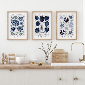 May include: Three framed prints with the title "Flower Market" and the city names "Paris", "Tokyo", and "New York" underneath. Each print features a different stylized floral design in shades of blue and green.