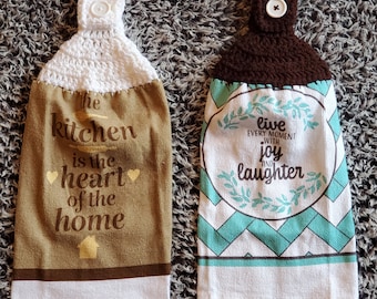 May include: Two brown and white kitchen towels with crocheted loops. The first towel has the text "The kitchen is the heart of the home" with a small house icon. The second towel has the text "Live every moment with joy and laughter" with a green wreath design.