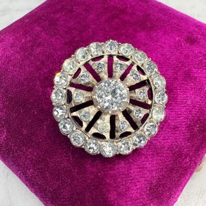 Peut inclure: Broche argentée de forme circulaire, ornée d'une grosse pierre transparente centrale entourée de petites pierres transparentes. La broche est posée sur un tissu de velours magenta foncé.