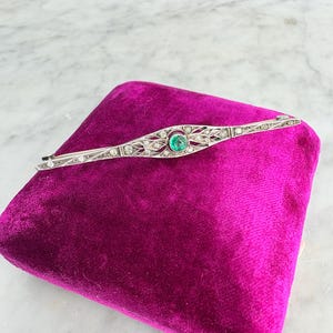 Peut inclure: Épingle à barre en argent avec une pierre centrale de couleur émeraude et des pierres claires environnantes. L'épingle est posée sur une surface de velours magenta. L'épingle a un design Art Déco.