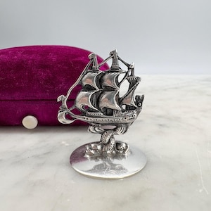 May include: A silver-toned miniature ship figurine with detailed sails and rigging, mounted on a round base. The ship is set against a blurred background, with a purple velvet box visible. The figurine is likely a decorative item or collectible.
