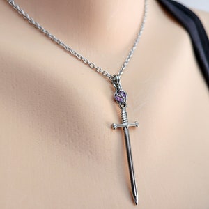 May include: A silver chain necklace with a sword pendant. The sword has a purple gemstone at the top.