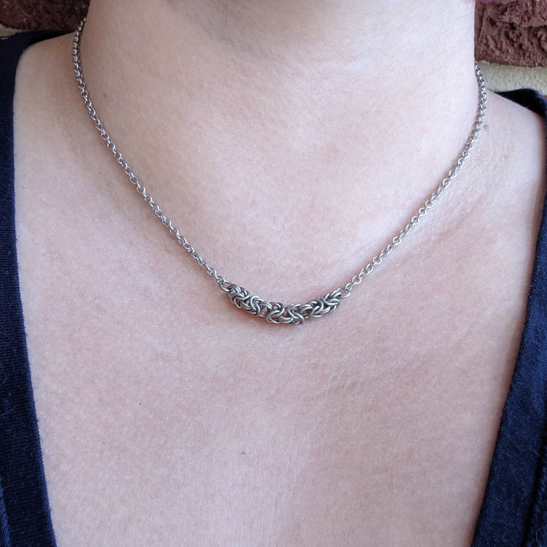 May include: A silver chain necklace featuring a distinctive, woven pendant. The pendant is crafted from numerous interlocked rings, resulting in a textured and intricate design.