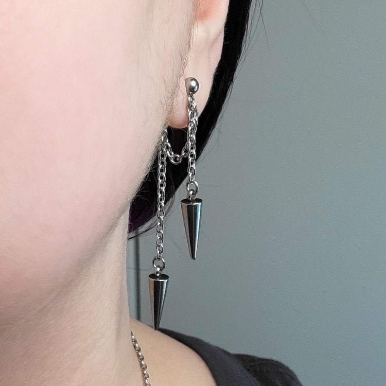 K&ouml;nnte beinhalten: Silberne Kettenohrringe mit zwei spitzen Metallspitzen, die an der Kette h&auml;ngen.