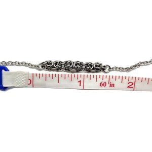 May include: A silver chain bracelet with a woven design, measured with a tape measure to be 1 1/2 inches long.