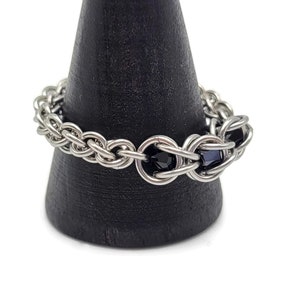 May include: A silver chain ring with black beads. The ring is on a black display stand.