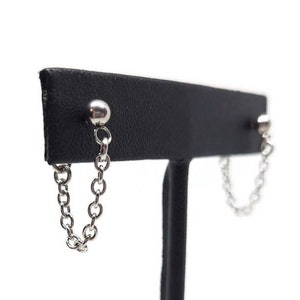 May include: A pair of silver chain earrings with small ball ends. The earrings are displayed on a black stand.