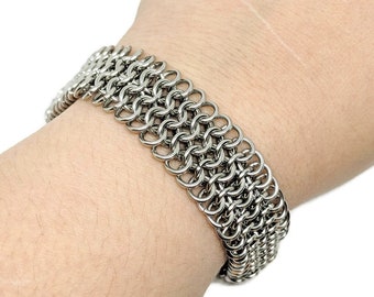 May include: A silver chainmail bracelet with a woven pattern. The bracelet is made of small, interlocking metal rings.