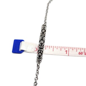 May include: A silver chain necklace with a woven metal design. The chain is measured against a ruler, showing it is about 1 inch long.