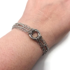 May include: A silver bracelet with multiple fine chain strands, joined to a central circular element. The bracelet, made of shiny metal, is shown on a wrist.