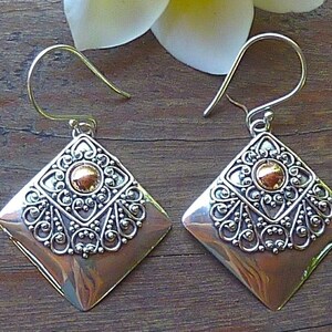 May include: A pair of diamond-shaped earrings with a copper-colored base and ornate silver filigree detailing. Each earring features a gold-colored central accent and a hook closure. The earrings are displayed on a wooden surface.