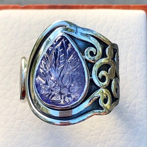 May include: A silver ring with a purple gemstone set in a teardrop shape. The ring has a decorative, scroll-like design around the gemstone.