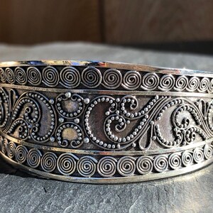 K&ouml;nnte beinhalten: Ein silbernes Armband mit komplizierten, wirbelnden Designs. Das Armband besteht aus einem dicken Metallband und hat eine polierte Oberfl&auml;che.