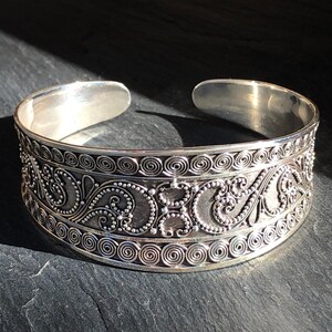 K&ouml;nnte beinhalten: Ein silbernes Armreif-Armband mit einem komplizierten, verzierten Design mit Spiralen und Schn&ouml;rkeln. Das Armband ist offen und verstellbar.
