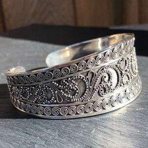 K&ouml;nnte beinhalten: Ein silbernes Armreif mit einem komplizierten Design aus Spiralen und Punkten. Das Armband ist breit und hat eine abgerundete Form.