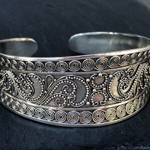 K&ouml;nnte beinhalten: Silberner Armreif mit einem dekorativen Design aus Spiralen und Punkten. Das Armband ist offen und verstellbar.