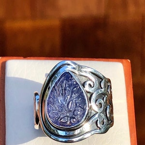 May include: A silver ring with an intricate design featuring a purple gemstone. The gemstone is set in a teardrop shape and has a carved floral pattern.