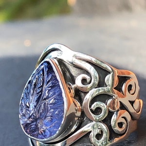 May include: A silver ring with a purple gemstone set in a teardrop shape. The ring has a detailed, ornate design with swirling patterns.