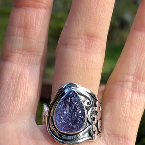 May include: A silver ring with an ornate design features a purple amethyst gemstone in a teardrop shape.