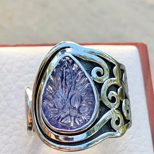 May include: A silver ring with a purple gemstone set in a floral design. The ring is wide and has a unique, curved shape.