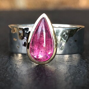 K&ouml;nnte beinhalten: Ein Ring mit einem tropfenf&ouml;rmigen rosafarbenen Edelstein, der in einer goldenen Fassung sitzt. Der Ringband ist aus Silber und hat eine geh&auml;mmerte Textur.