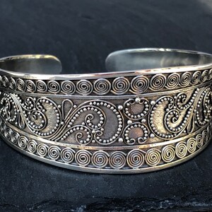 K&ouml;nnte beinhalten: Ein silbernes Armreif mit einem komplizierten Design aus Spiralen und Punkten. Das Armband ist aus Metall und hat eine polierte Oberfl&auml;che.