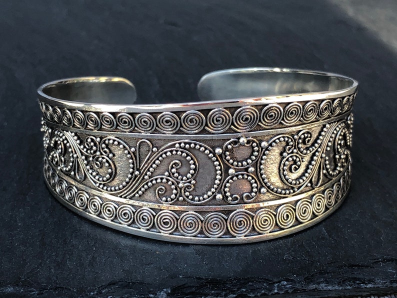 K&ouml;nnte beinhalten: Ein silbernes Armreif mit einem komplizierten Design aus Spiralen und Punkten. Das Armband ist aus Metall und hat eine polierte Oberfl&auml;che.