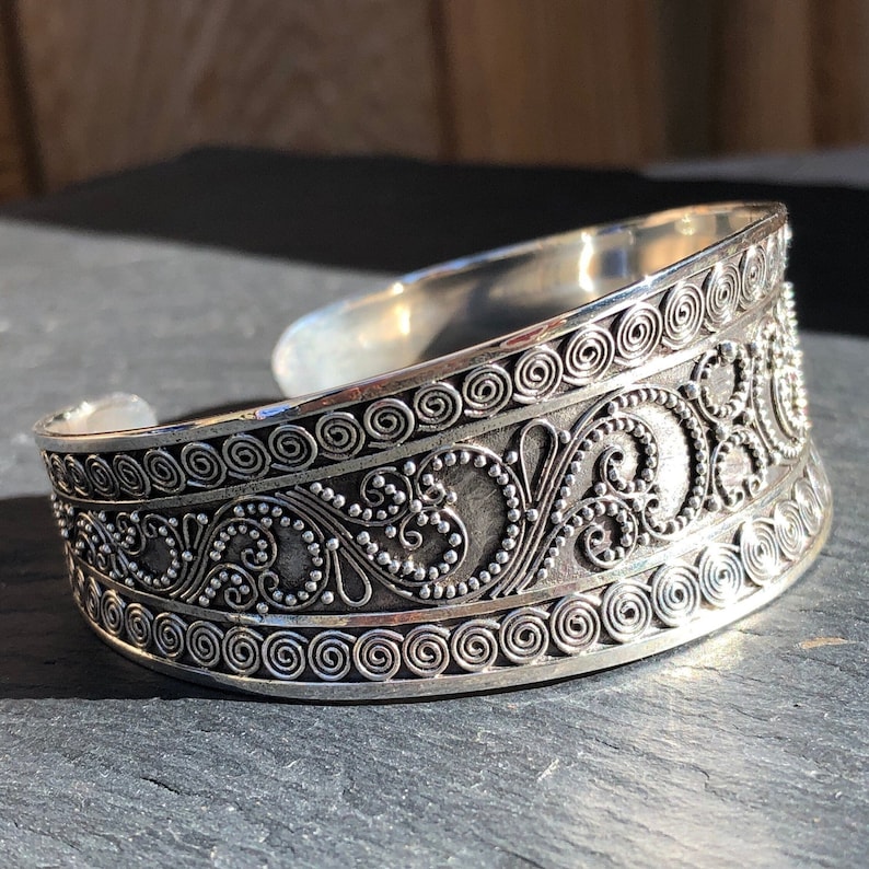K&ouml;nnte beinhalten: Ein silbernes Armreif mit einem komplizierten Design aus Spiralen und Punkten. Das Armband ist breit und hat eine abgerundete Form.