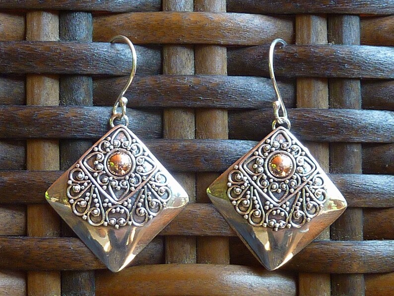 May include: A pair of silver-toned earrings with a diamond shape. Each earring features a central copper-colored stone surrounded by ornate silver filigree. The earrings hang from a simple hook.