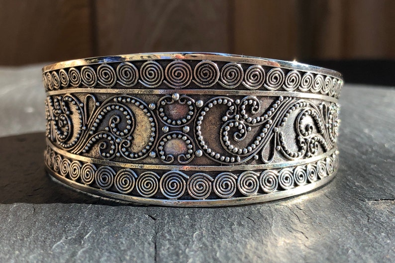 K&ouml;nnte beinhalten: Silberarmband mit aufwendigen floralen und spiralf&ouml;rmigen Designs. Das Armband ist breit und hat eine polierte Oberfl&auml;che.