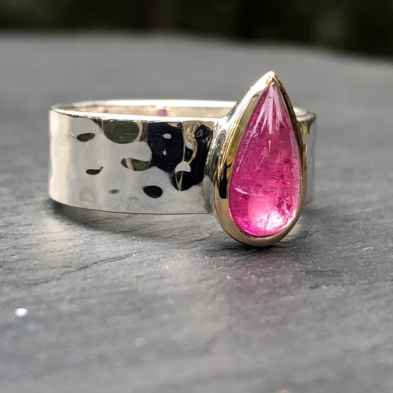K&ouml;nnte beinhalten: Ein silberner Ring mit geh&auml;mmertem Muster und einem tropfenf&ouml;rmigen, pinkfarbenen Edelstein, der in einer goldenen L&uuml;nette gefasst ist. Das Ringband ist breit und hat eine strukturierte, geh&auml;mmert Oberfl&auml;che.