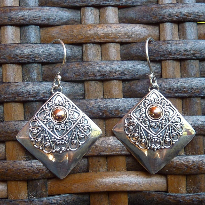 May include: A pair of silver-toned, diamond-shaped earrings with intricate filigree detailing. Each earring features a central, circular, copper-colored accent. The earrings have a hook closure and are displayed against a woven background.