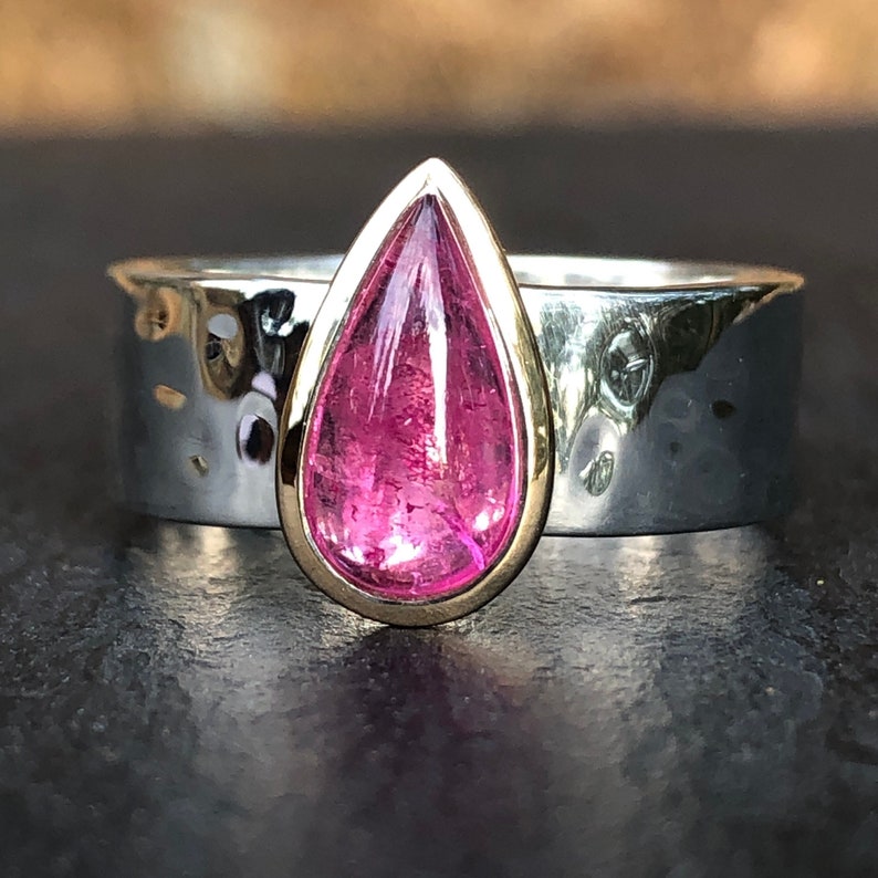 K&ouml;nnte beinhalten: Ein Ring mit einem tropfenf&ouml;rmigen rosafarbenen Edelstein, der in einer goldenen Fassung sitzt. Der Ringband ist aus Silber und hat eine geh&auml;mmerte Textur.